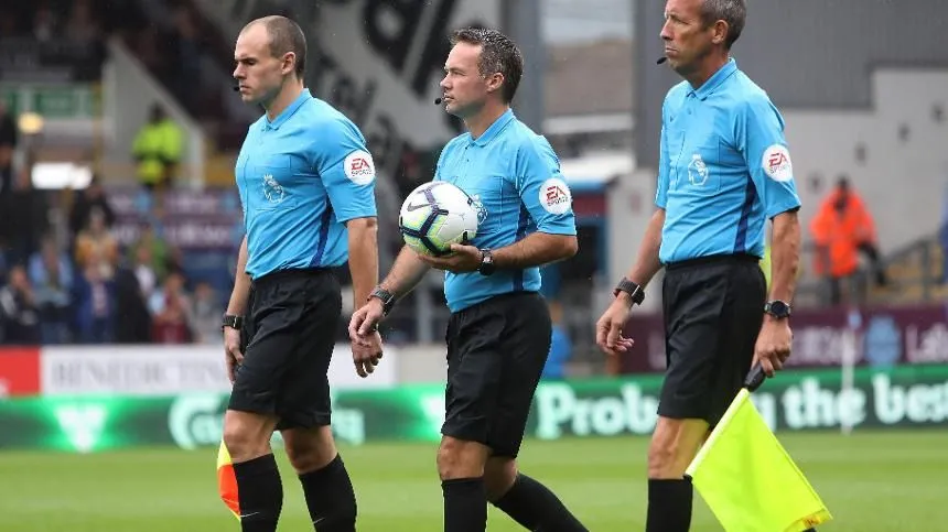 three referees wearing blue shirts and black shorts walking on a football pitch while the middle one is holding a ball