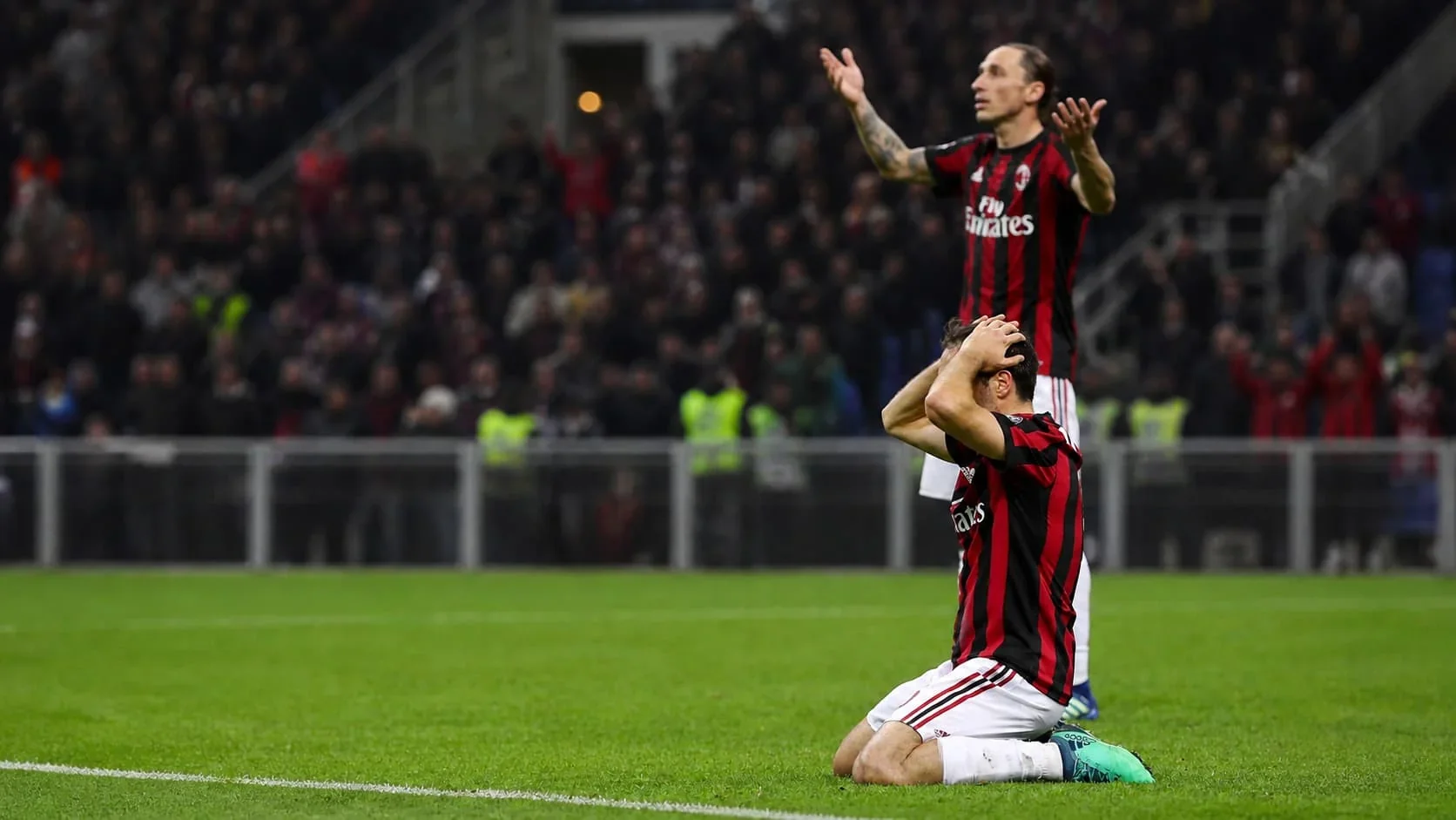 Two AC Milan players react in frustration on the pitch, one kneeling with his hands on his head and another standing behind him in a blurred stadium crowd.