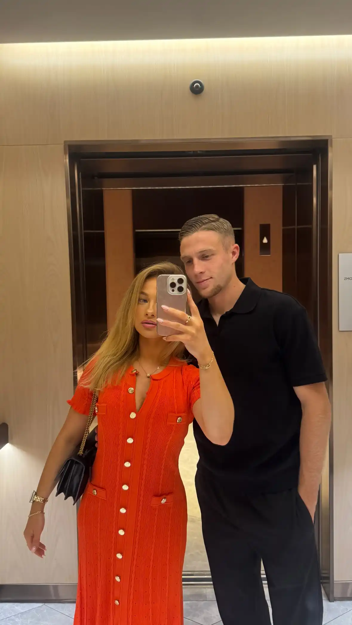 floraine allain and adrien truffert selfie in the elevator in a hotel she wears orange dress and he is wearing all black polo tshirt and black trousers