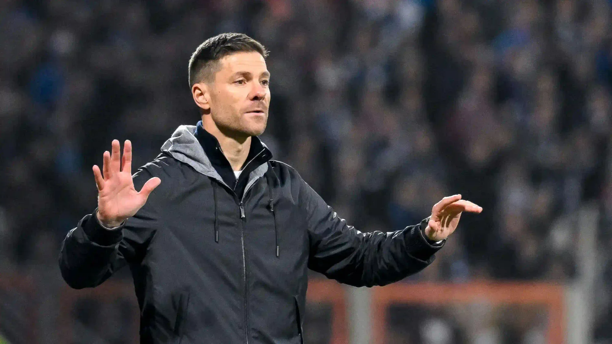 Xabi Alosno in a dark jacket with a grey hood, gesturing from the touchline with a blurred stadium crowd behind.