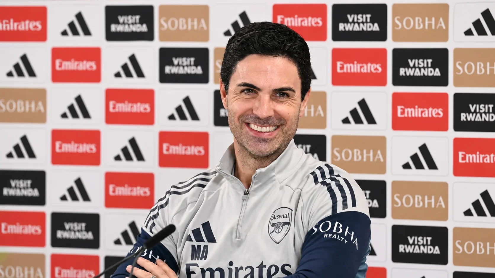 Arteta smiling at a press conference in Arsenal training gear with a sponsor backdrop