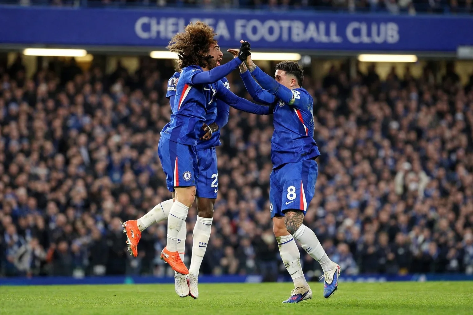 Three Chelsea players in blue kits celebrate with a mid-air high-five on the pitch, with the “Chelsea Football Club” stand signage in the background.