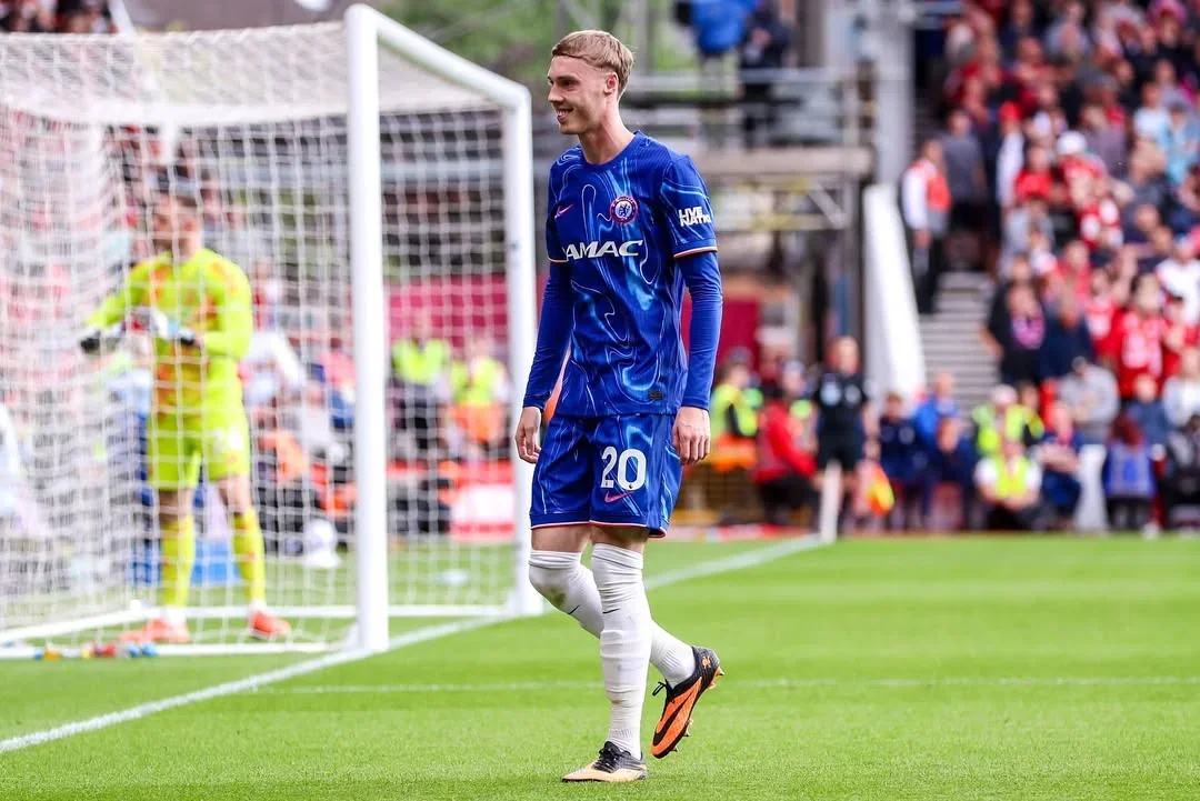 Cole Palmer walking on the pitch in a Chelsea blue kit during a match, with the goal and crowd in the background.