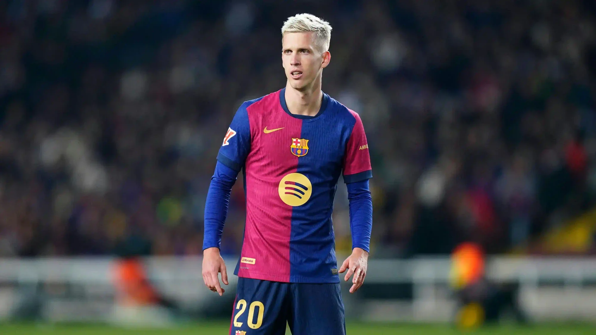 Dani Olmo wearing a Barcelona home kit during a match, standing on the pitch with the crowd blurred in the background.