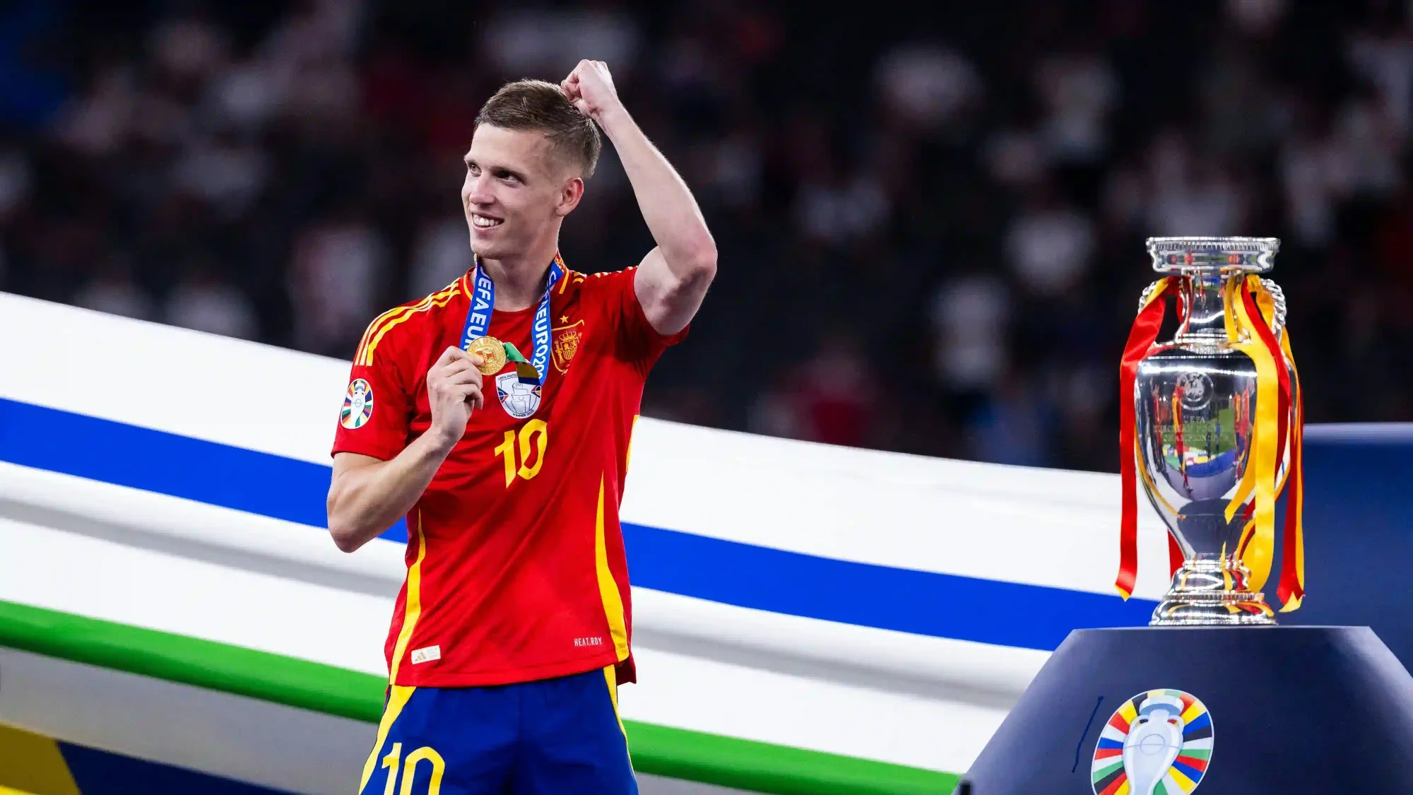 Dani Olmo in a Spain kit holding up his winner’s medal on the podium next to the tournament trophy.