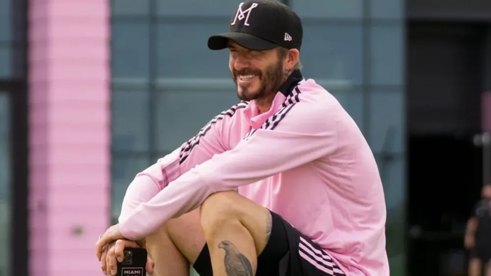 david beckham sitting on a ball wearing inter miami pink shirt and smiling