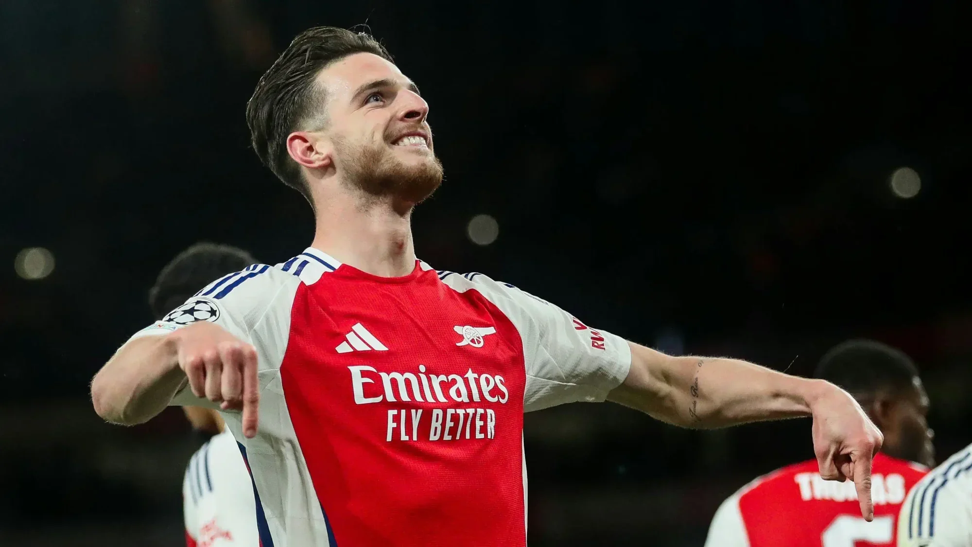 Declan Rice in an Arsenal home kit celebrates with arms outstretched during a match, smiling and looking upward under stadium lights.