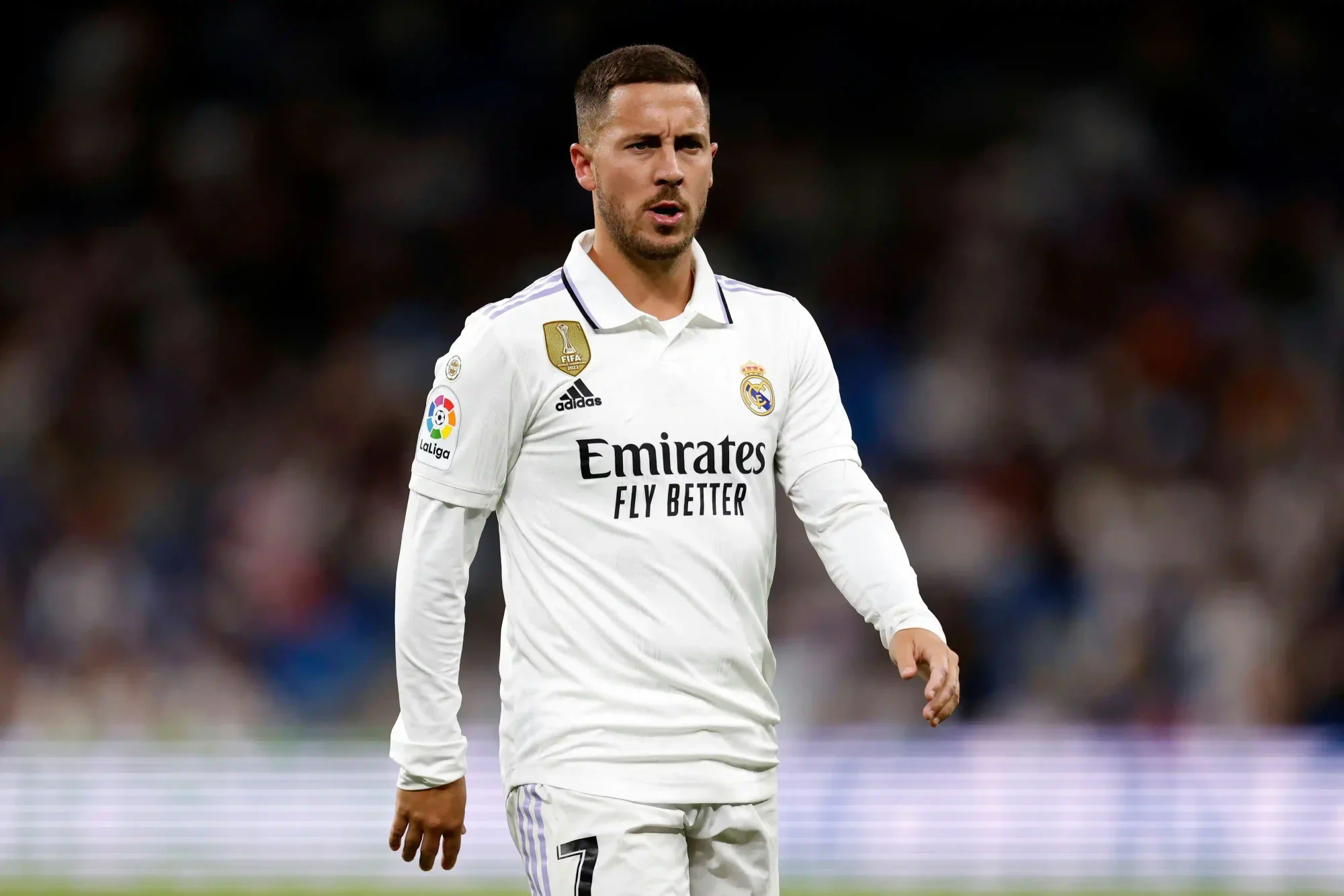 Eden Hazard in a white Real Madrid kit walking on the pitch during a match, with a blurred stadium crowd in the background  Select 77 more words to run Humanizer.