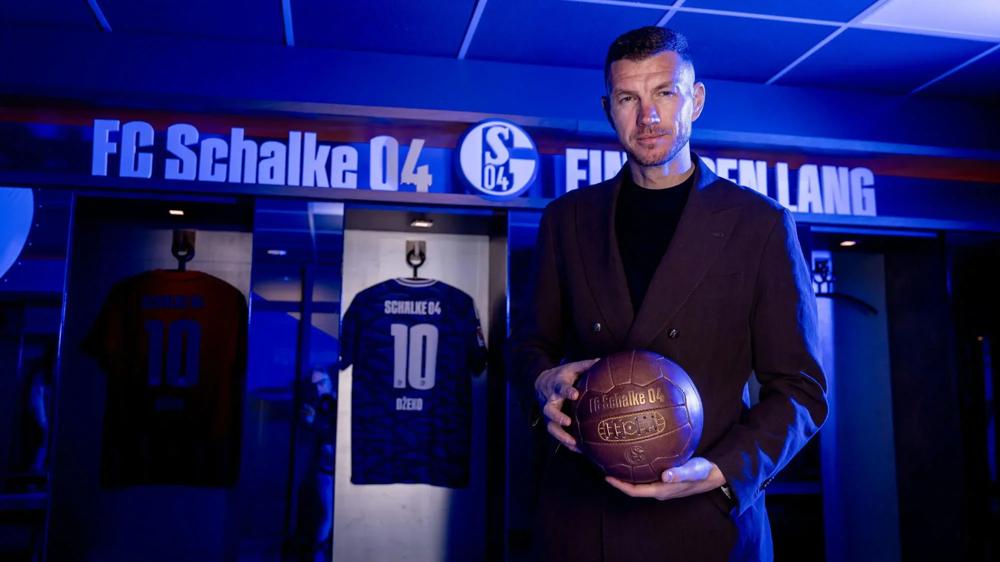 Edin Džeko holding a football inside an FC Schalke 04 stadium area, with a Schalke 04 shirt displayed in the background under blue lighting.