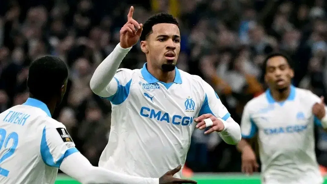 Ethan Nwaneri celebrating a goal in an Olympique de Marseille white and light-blue kit during a match, with teammates running toward him.