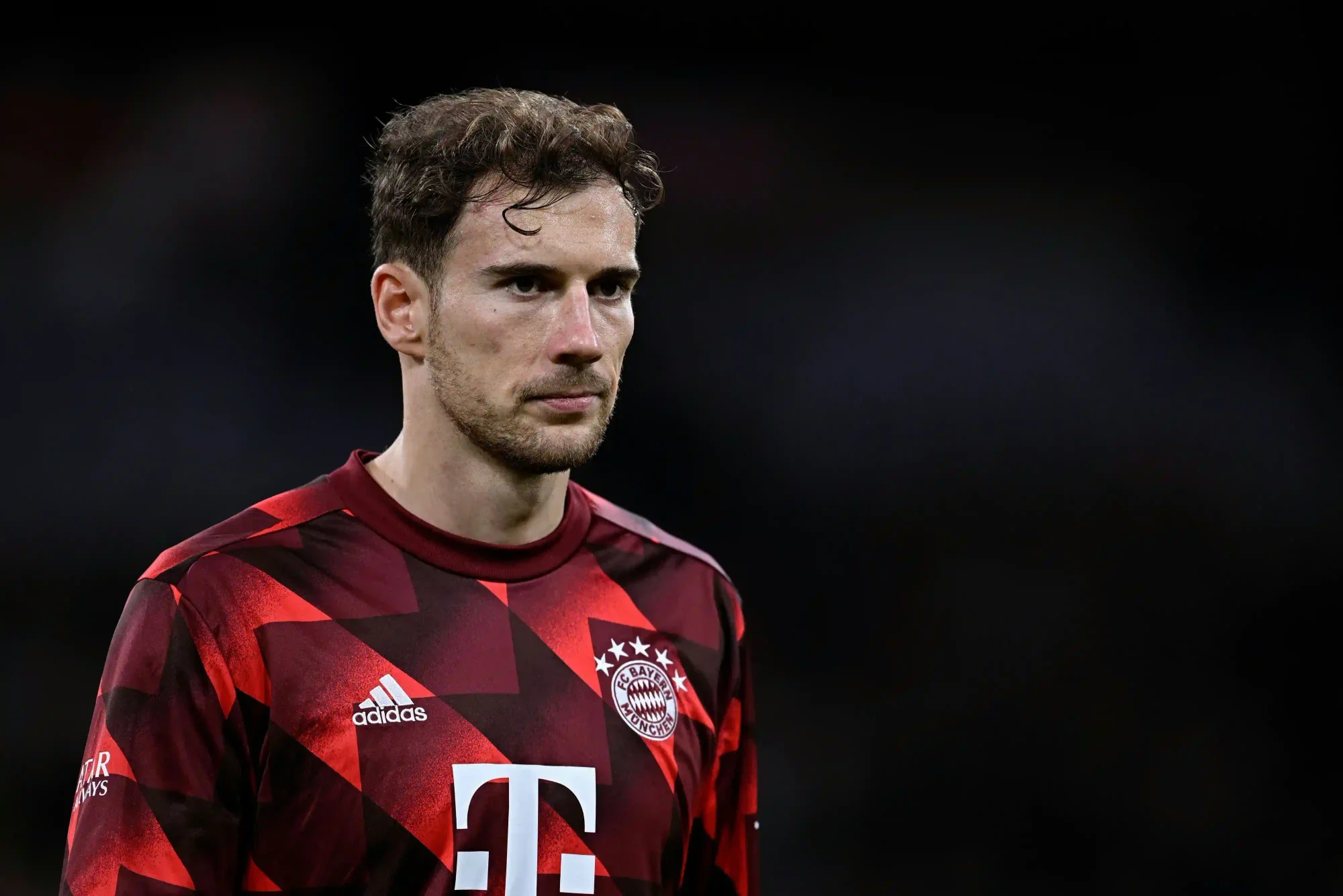 Close-up portrait of a Bayern Munich Leon Goretzka wearing a red-and-black patterned jersey, looking focused against a dark blurred stadium background.