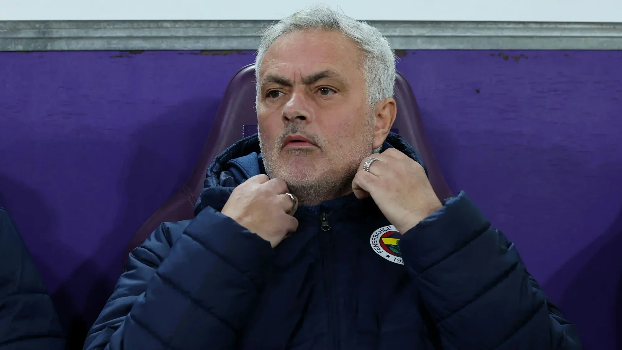 José Mourinho sits on the bench wearing a Fenerbahçe jacket during a match.