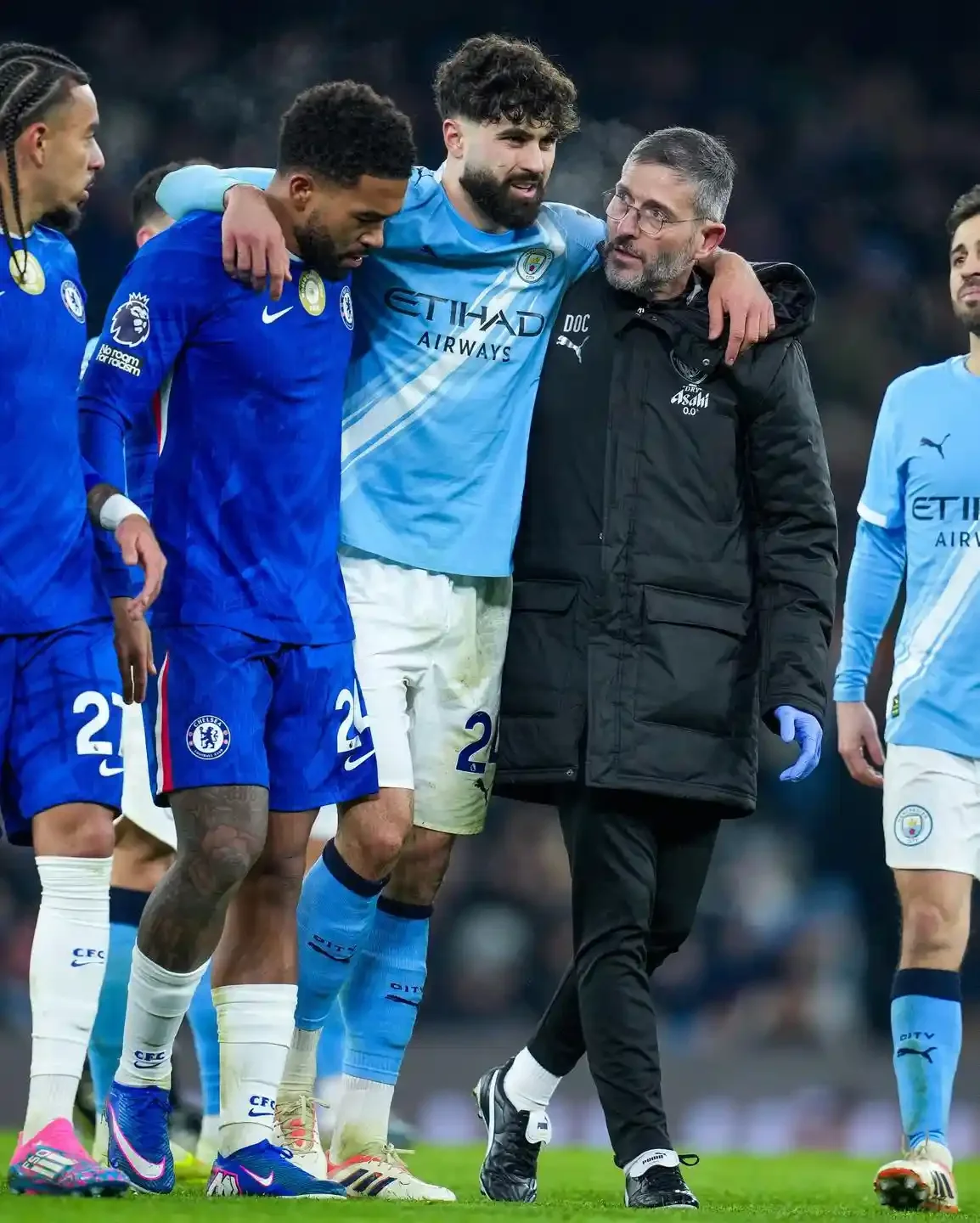 Josko Gvardiol is helped off the pitch by Manchester City staff and Chelsea player after picking up an injury during a match, with opposing players nearby.