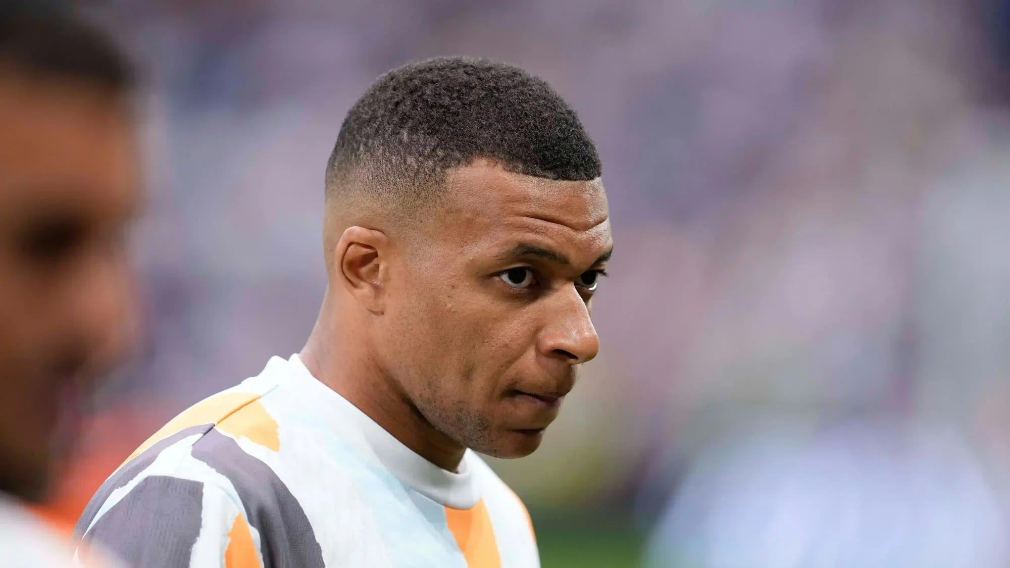 Close-up of Kylian Mbappé during warm-up, looking focused with a blurred stadium background.