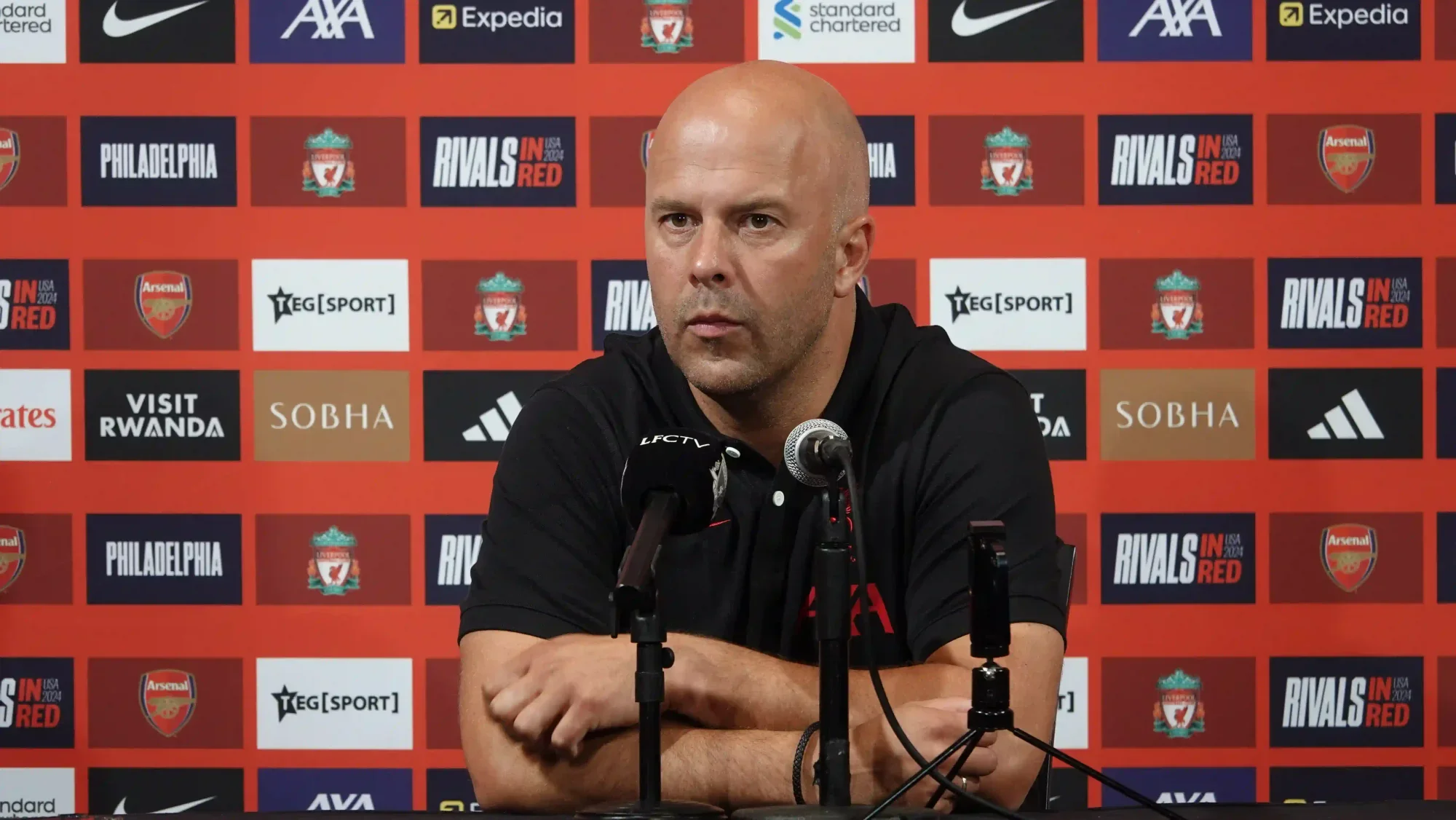 Liverpool manager Arne Slot speaking at a press conference, seated behind microphones with a red sponsor backdrop.