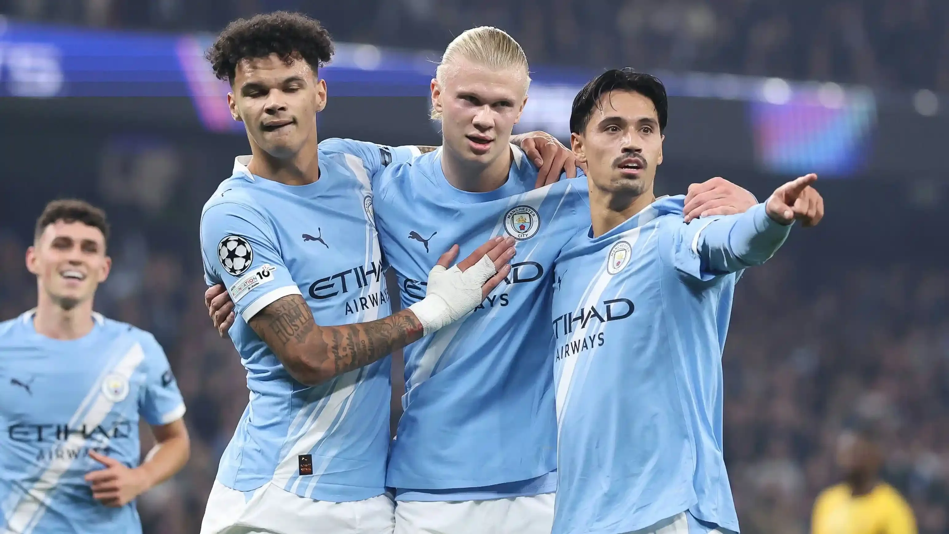 Three Manchester City players in sky-blue kits celebrate, with one pointing toward the stands.