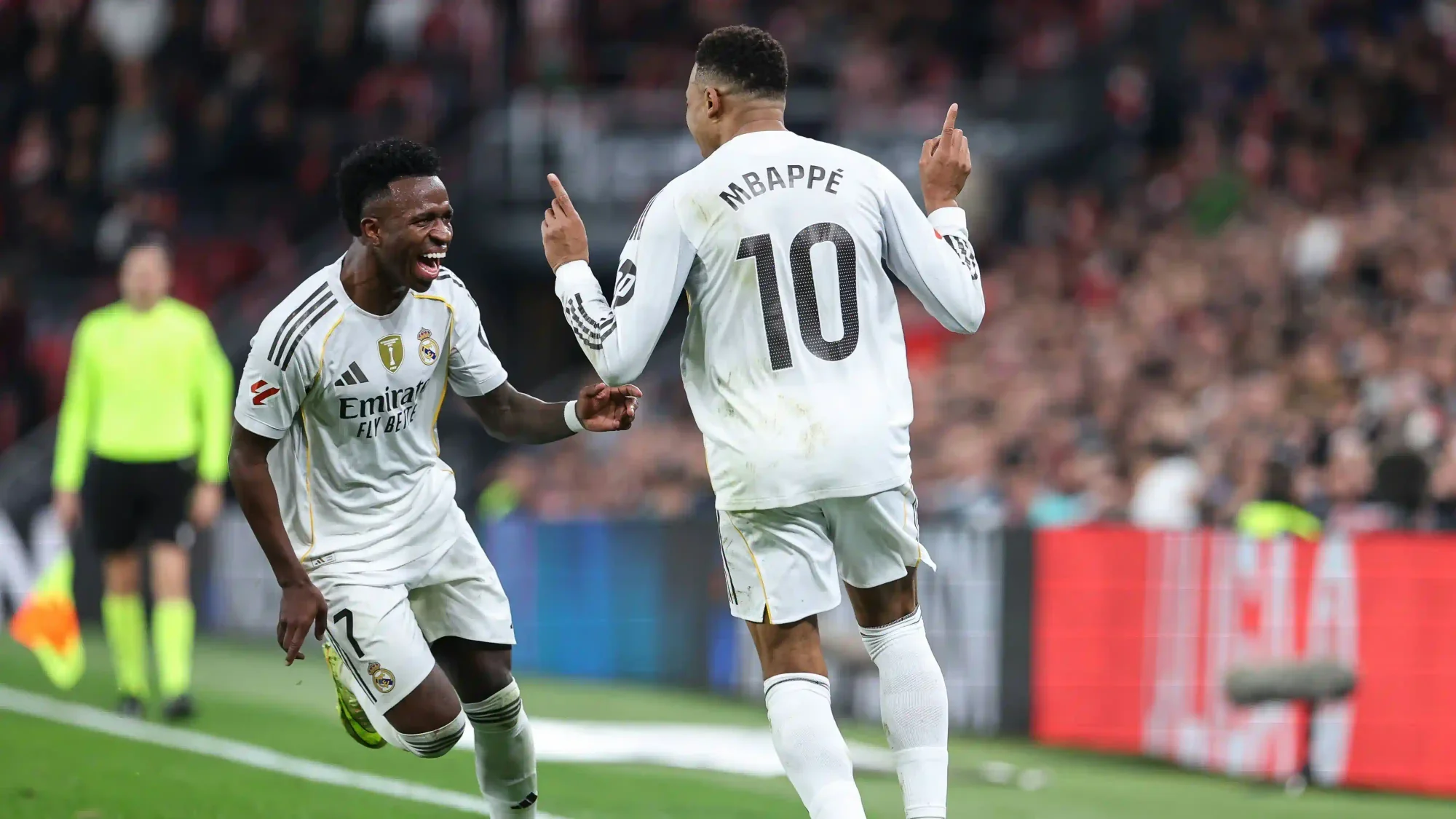 Kylian Mbappé and Vinícius Júnior celebrating together in Real Madrid’s white kit during a match, with a crowd in the background.