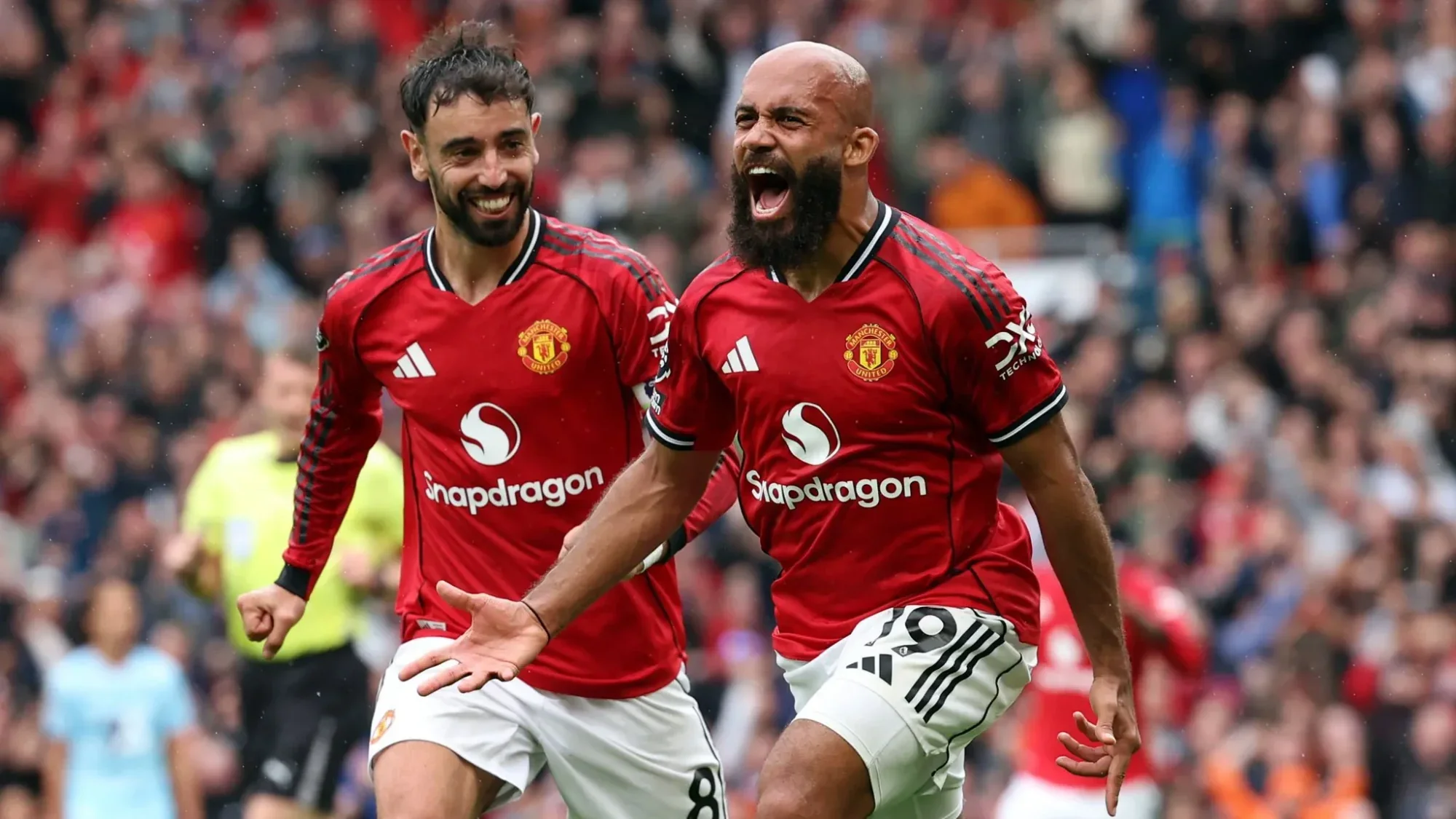 Bruno Fernandes and Bryan Mbuemo celebrate on the pitch, one shouting with arms outstretched while the other smiles as they run, with a blurred crowd in the backgroun