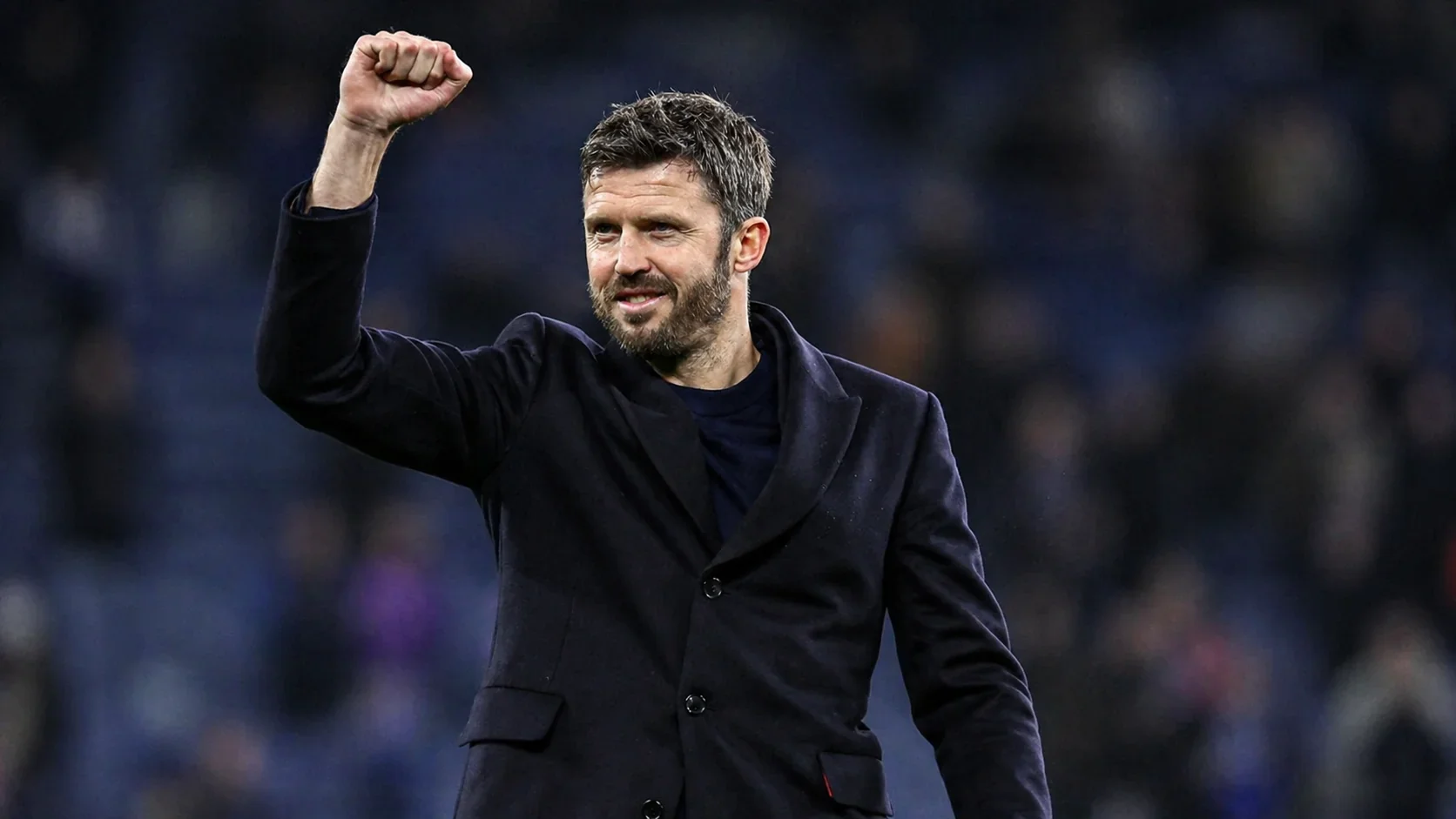 Michael Carrick raising his fist while wearing a dark coat on the touchline in a stadium
