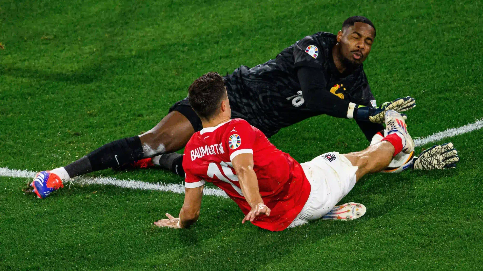 France goalkeeper Mike Maignan making a save on the ground during a match, with an opposing player sliding nearby.