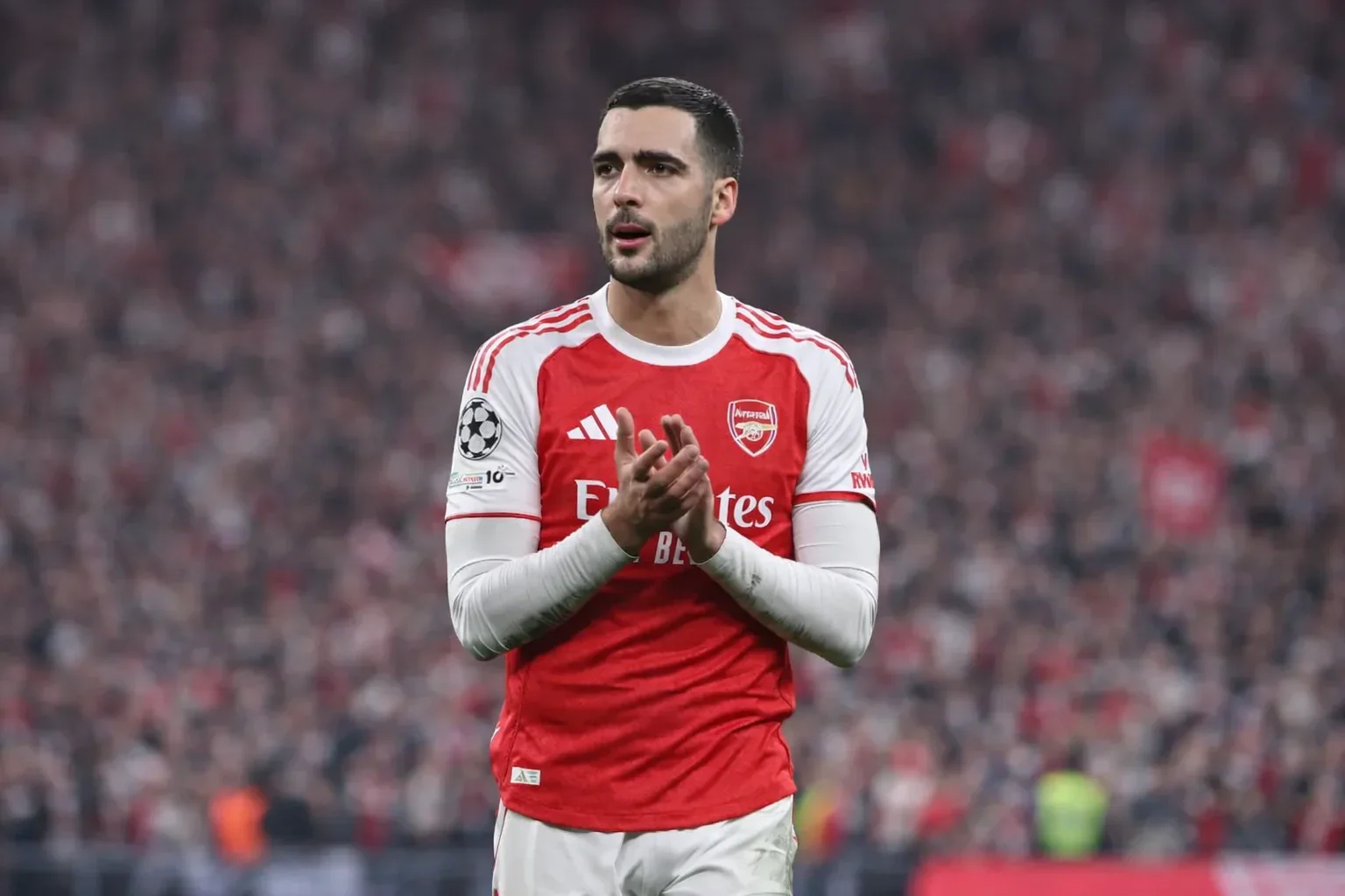 Mikel Merino applauding in an Arsenal home kit during a Champions League match, with a blurred stadium crowd in the background.