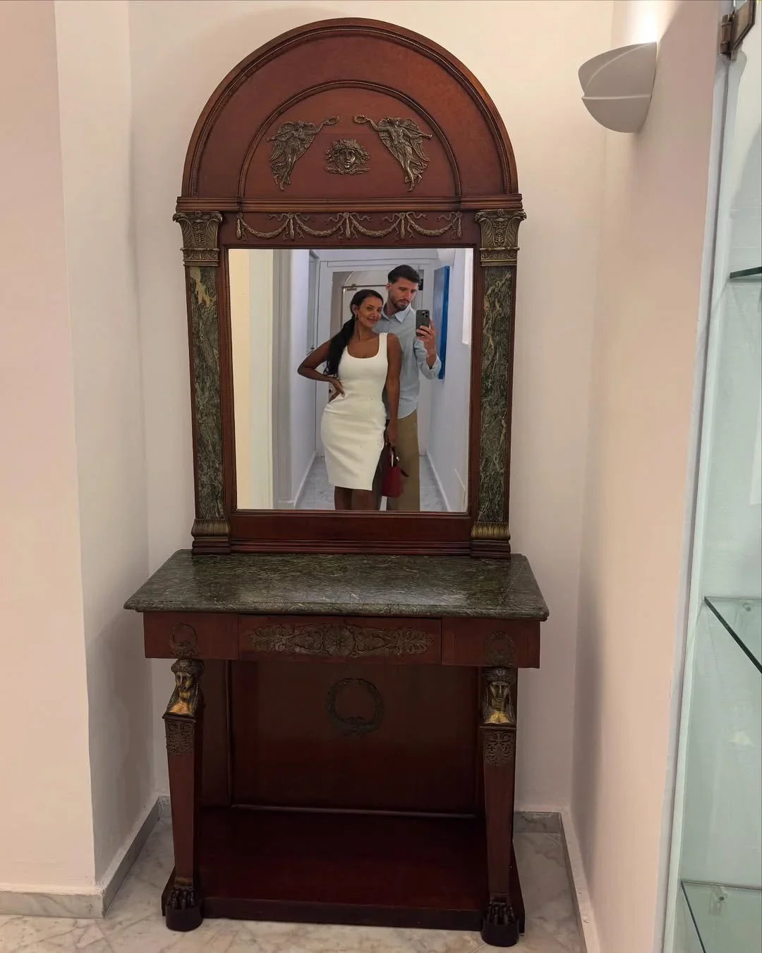 ruben dias and maya jama taking a mirror selfie in a hallway, maya jama in a white dress and dias in a light shirt standing behind her