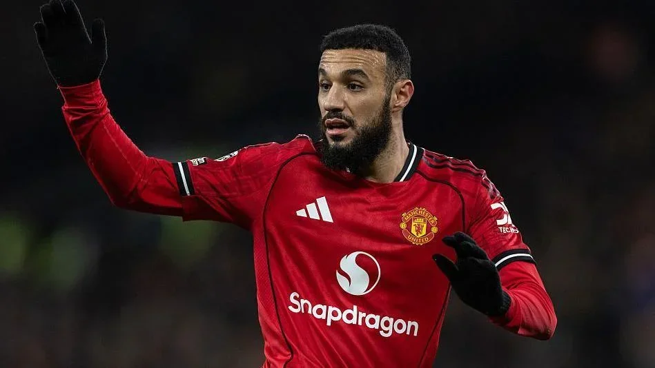 Manchester United player mazraoui in a red home kit raising his arm during a match, wearing black gloves with a dark blurred stadium background