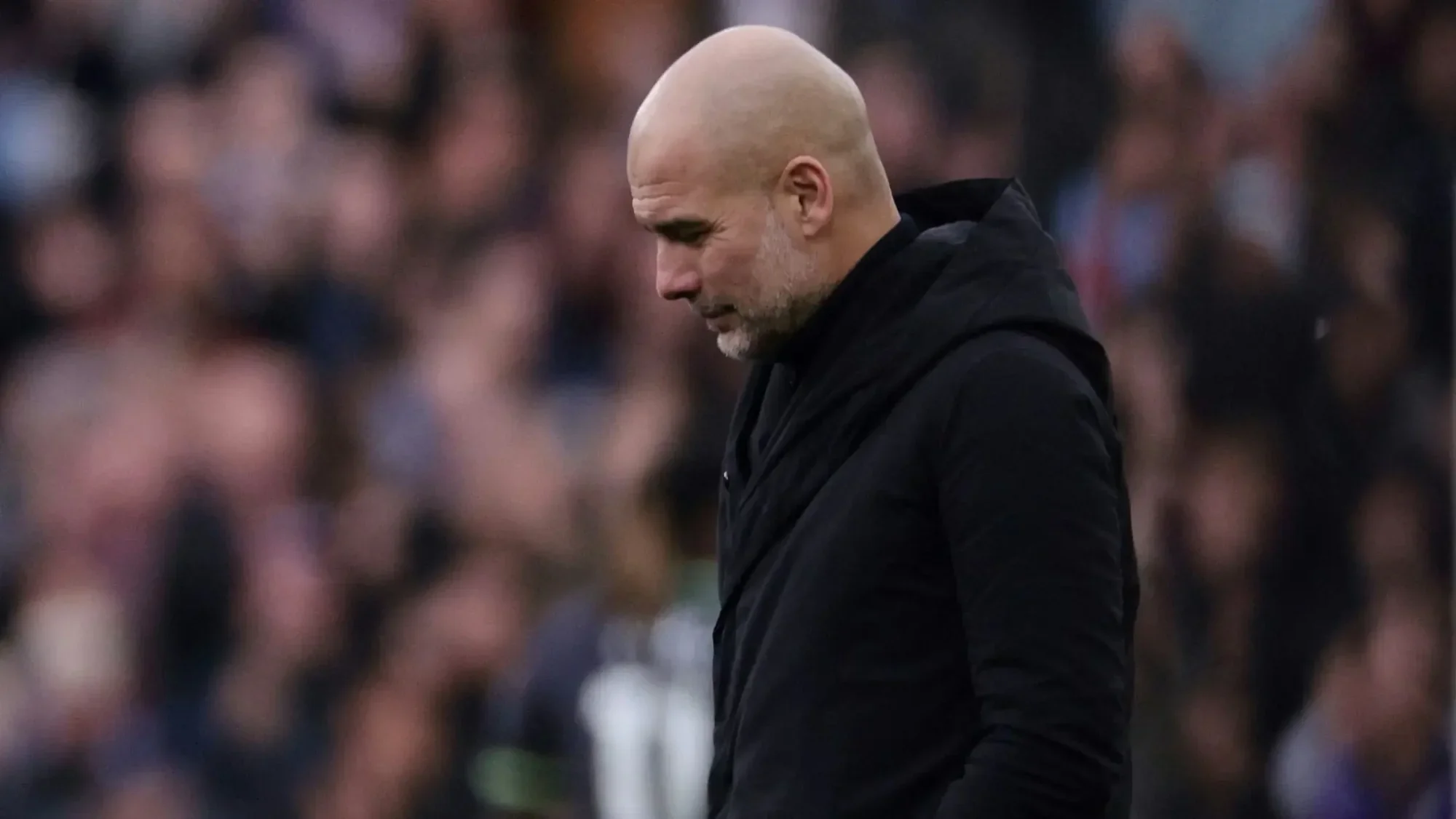 Pep Guardiola looks dejected on the touchline during a Manchester City match, with a blurred crowd in the background.