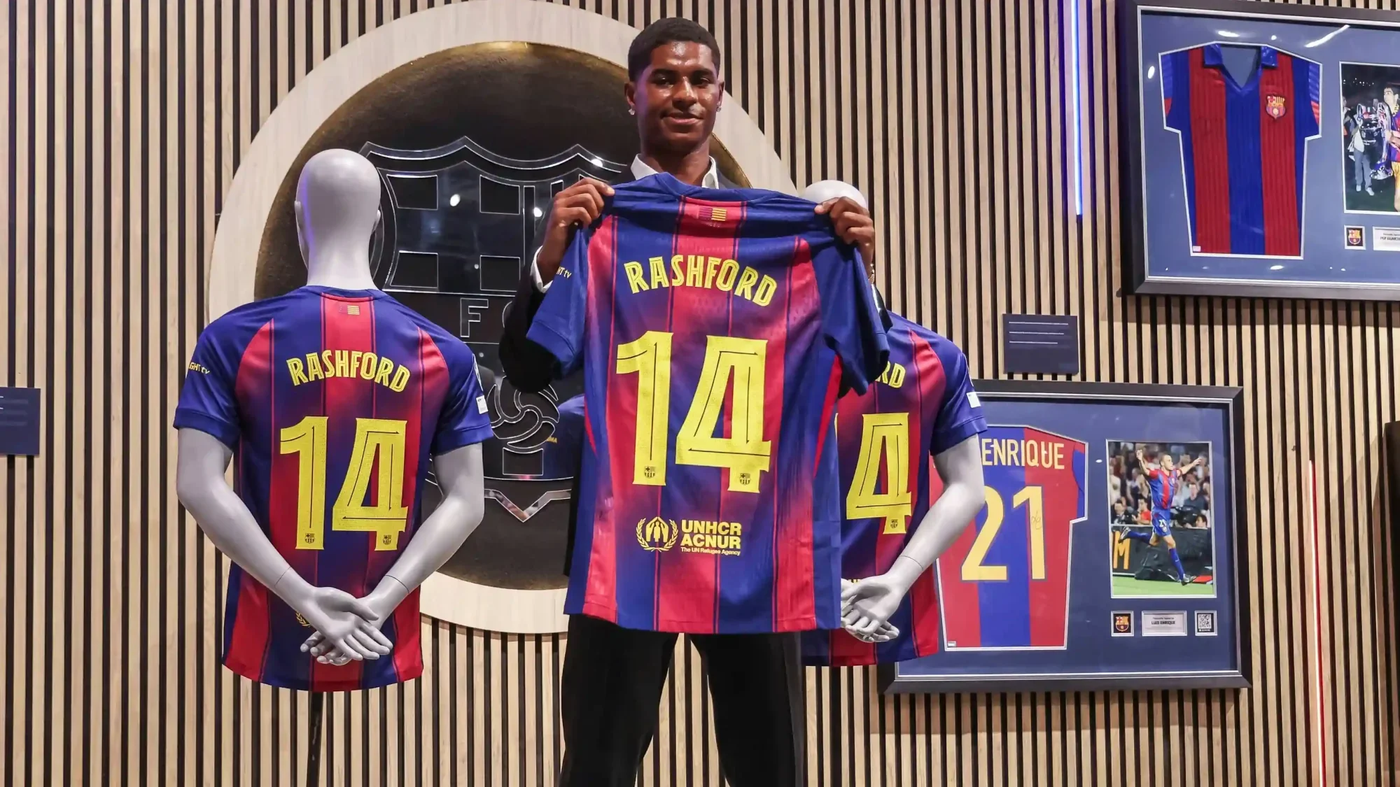 Rashford olding a Barcelona home shirt with “RASHFORD” and number 14 during a presentation, with matching shirts displayed on mannequins behind him.