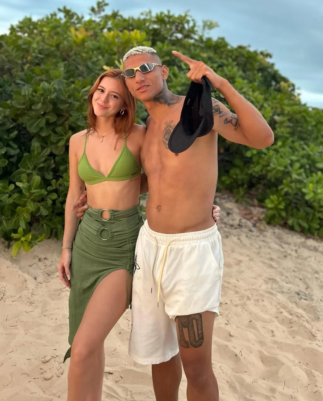 Amanda Araújo and Richarlison standing on a beach, Amanda in a green bikini and skirt, Richarlison shirtless in white shorts, smiling with his arm around her.