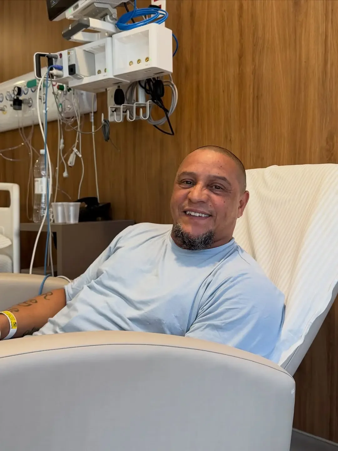 Roberto Carlos smiles while sitting in a hospital chair, with medical equipment behind him.
