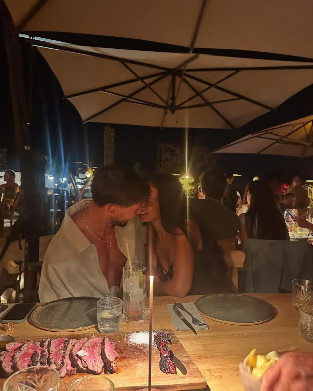 ruben diasn and maya jama sitting close together at a restaurant table at night with steak and drinks in the foreground