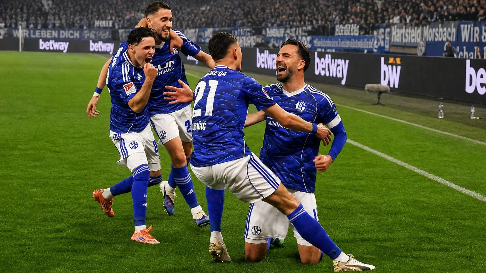 Schalke players celebrating a goal on the pitch in front of a packed stadium crowd