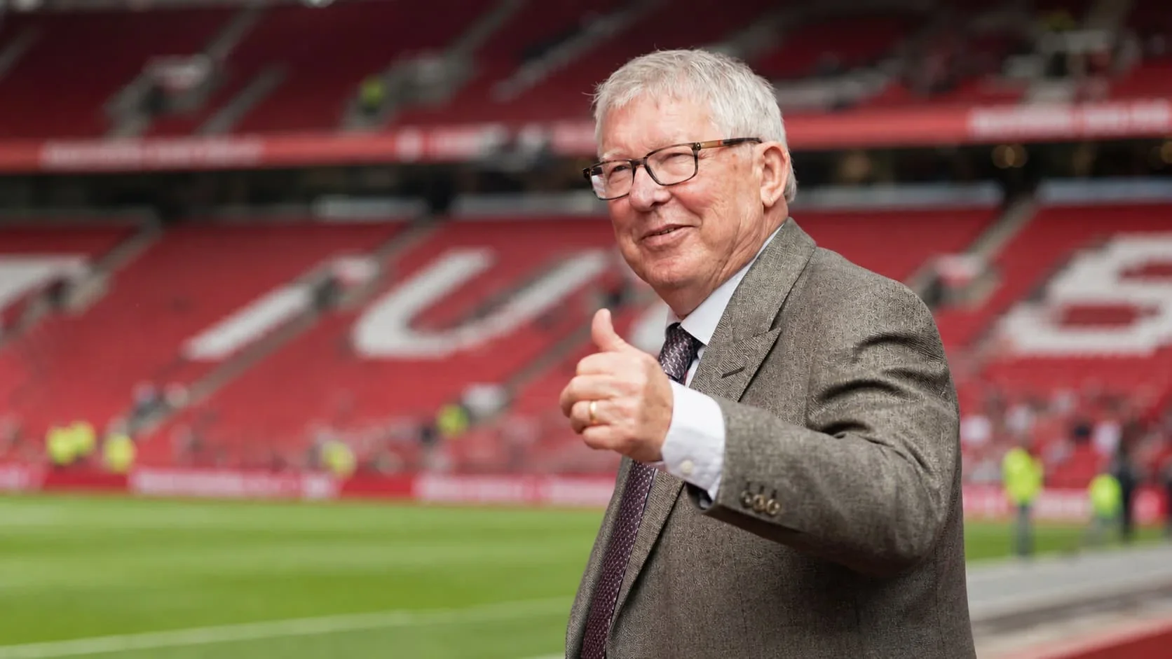 sir alex ferguson a grey suit and glasses gives a thumbs-up while standing beside the pitch in a football stadium with red seats in the background.