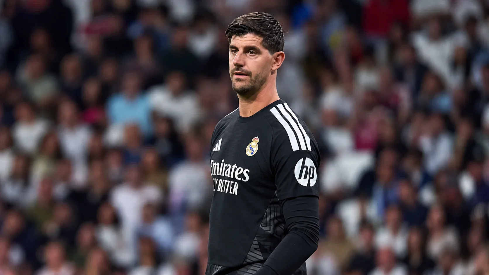 Thibaut Courtois in a black Real Madrid goalkeeper kit with a blurred stadium crowd behind him.