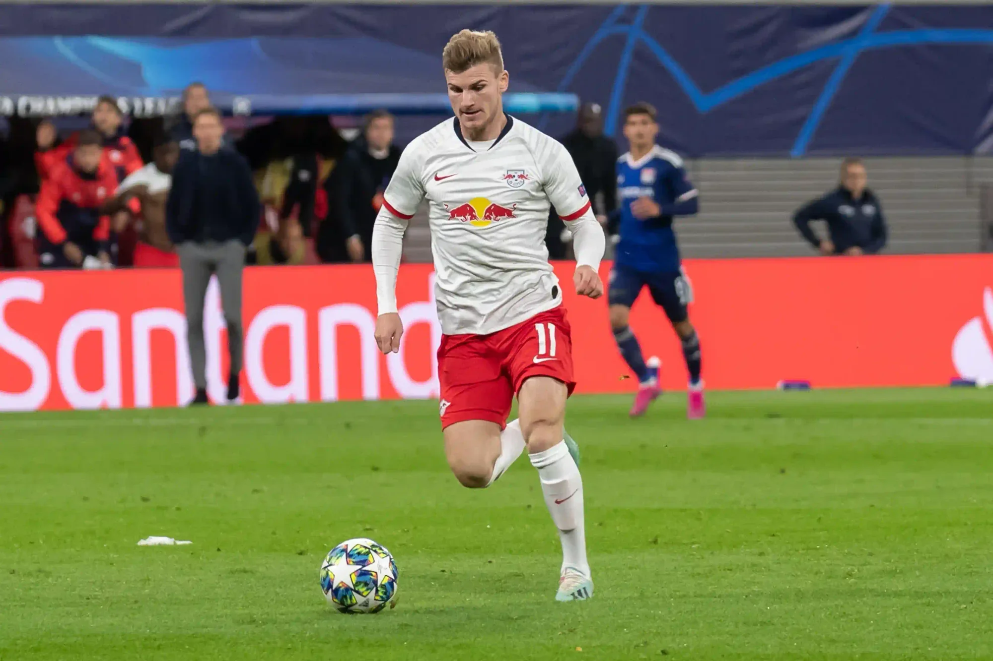 timo werner in a white shirt and red shorts dribbling the ball during a UEFA Champions League match, with the crowd and benches in the background.