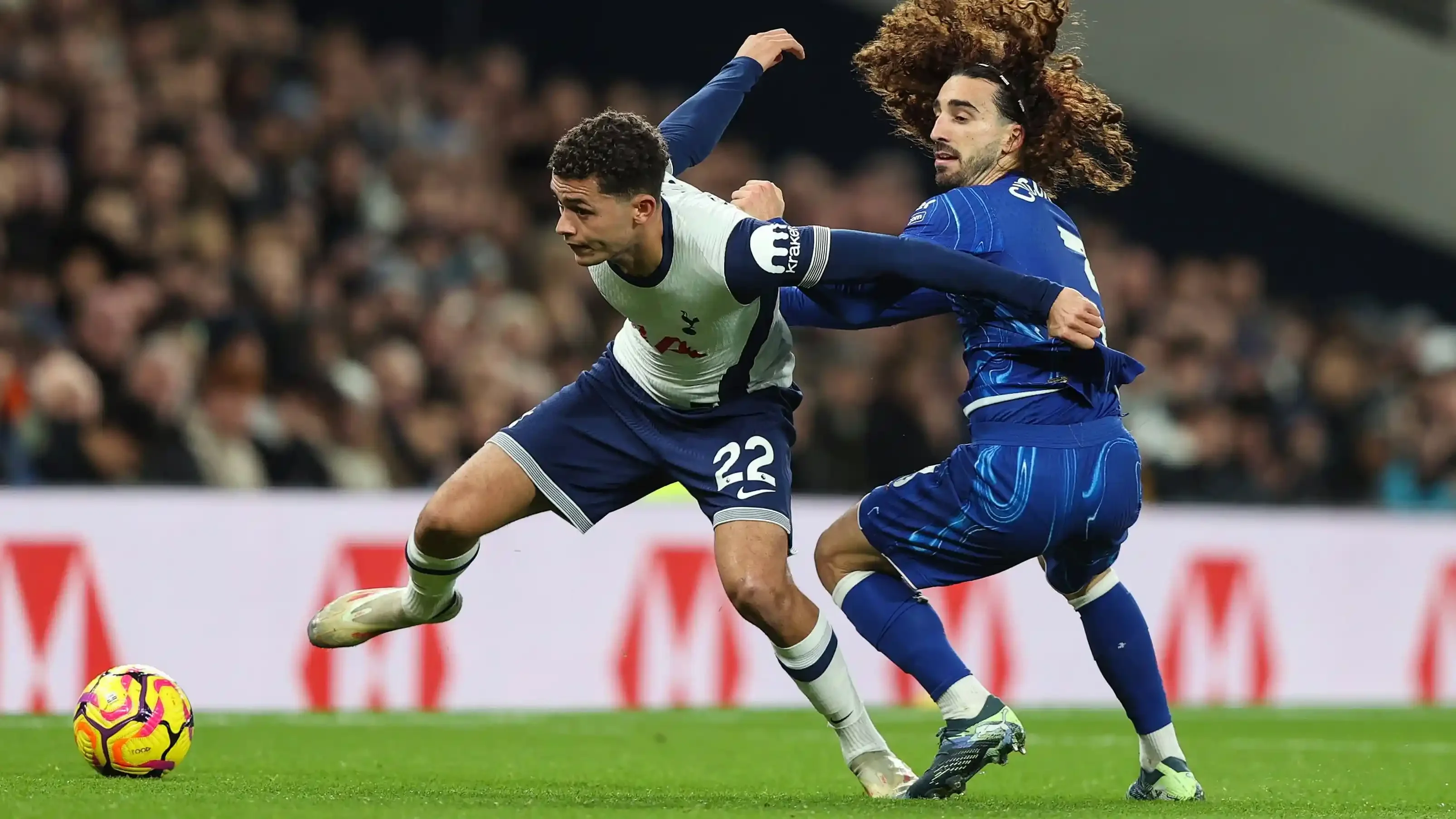 A Tottenham player shields the ball while a Chelsea player challenges from behind in a physical duel.