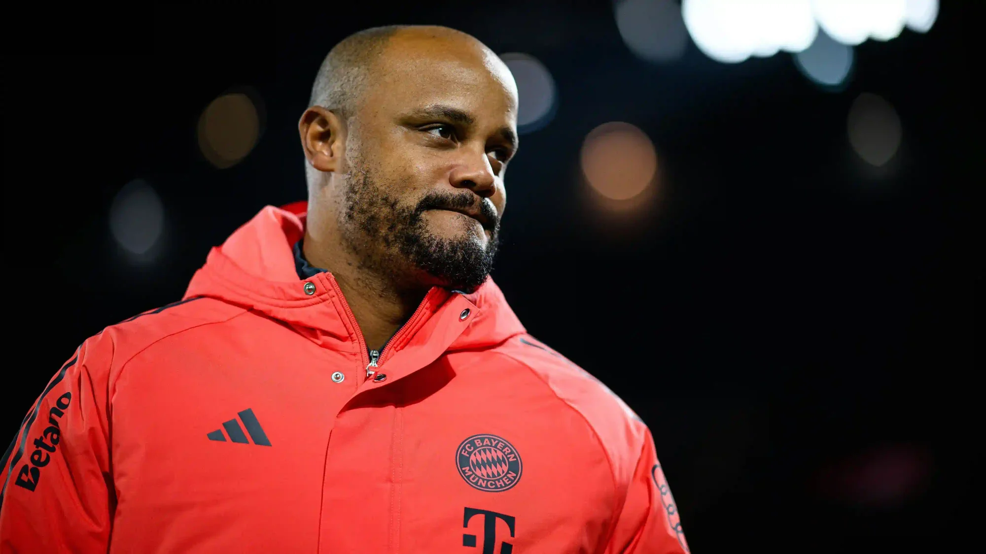 Vincent Kompany wearing a red Bayern Munich jacket, looking on before a match under stadium lights.