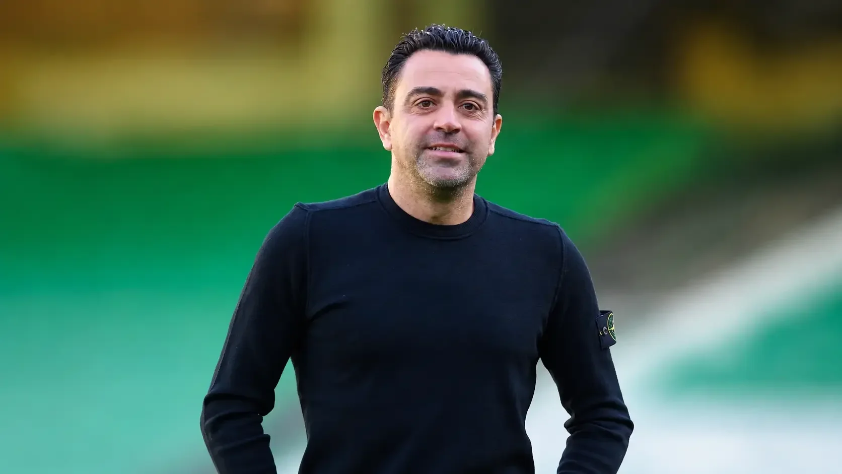 Xavi Hernández standing on the touchline in a black shirt